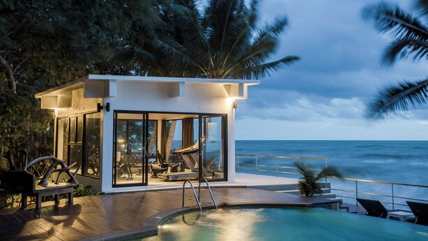 A modern outdoor pool area with a glass-enclosed gym, lounge chairs, and a view of the ocean.