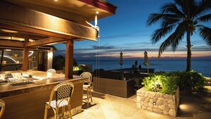 An outdoor bar area with a stone planter, wicker furniture, and a view of the ocean at dusk.
