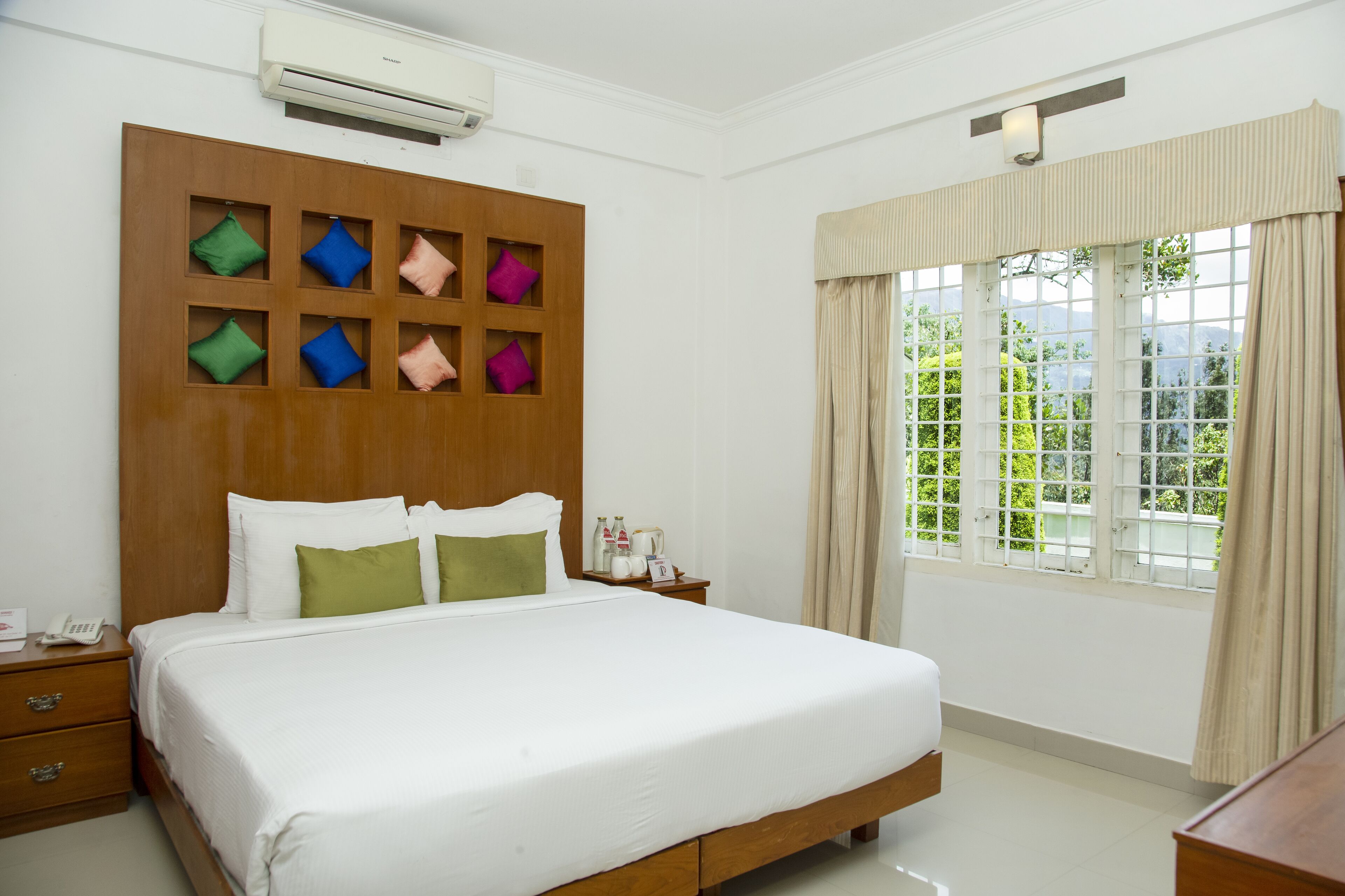 Habitación de hotel con una cama grande, cabecero de madera con compartimentos de almacenamiento, mesita de noche con teléfono y hervidor, y una ventana con cortinas.