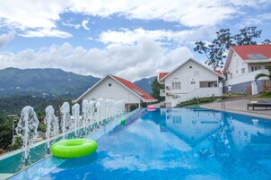 A swimming pool with a water fountain feature, surrounded by white buildings with red roofs, set against a mountainous backdrop.