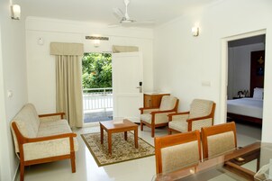 Habitación de hotel con sofá, sillas, una mesita, una mesa de centro de vidrio, cama y vistas a zonas verdes.
