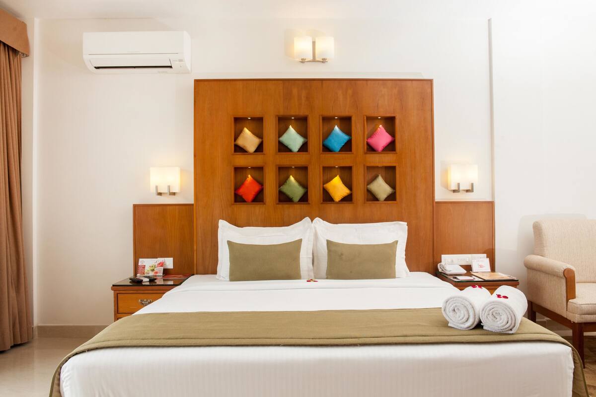 Ein Hotelzimmer mit einem großen Bett, einem hölzernen Kopfteil mit vielen Kissen, einem Nachttisch mit Telefon, einem Sessel und einer Klimaanlage an der Wand.