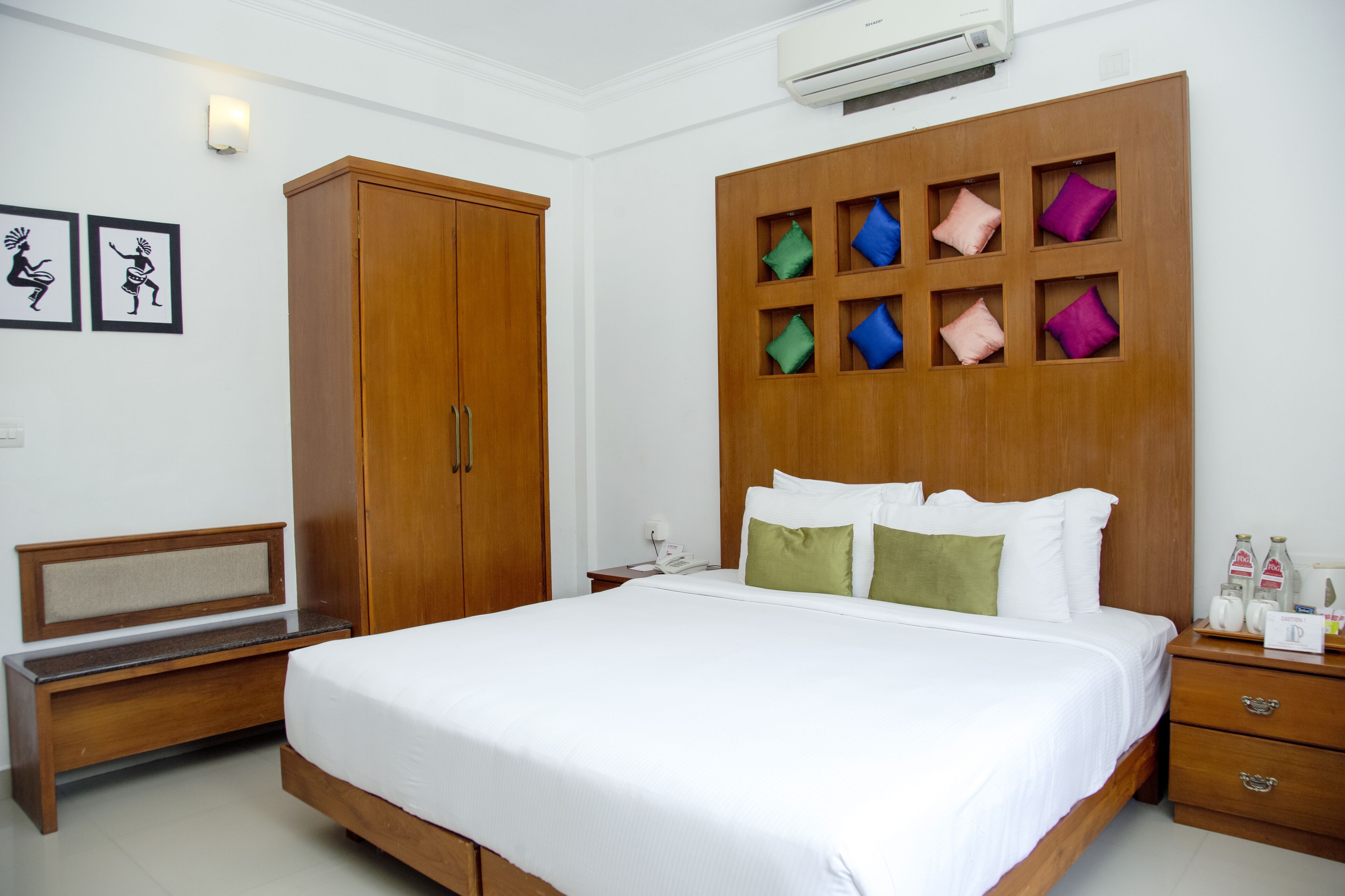 A hotel room with a large bed, a wooden wardrobe, a bench, and a wall-mounted shelf with colorful cushions.