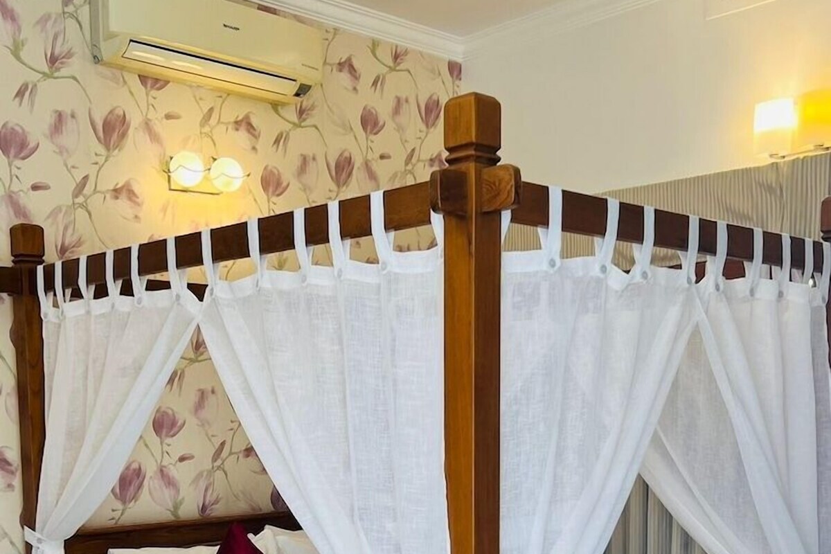 A four-poster bed with white curtains, a wooden headboard, and a floral wallpaper pattern.