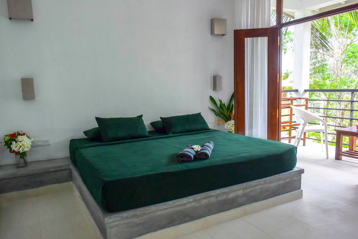 A bedroom with a bed, green bedding, a small table with chairs, and a view of greenery outside.