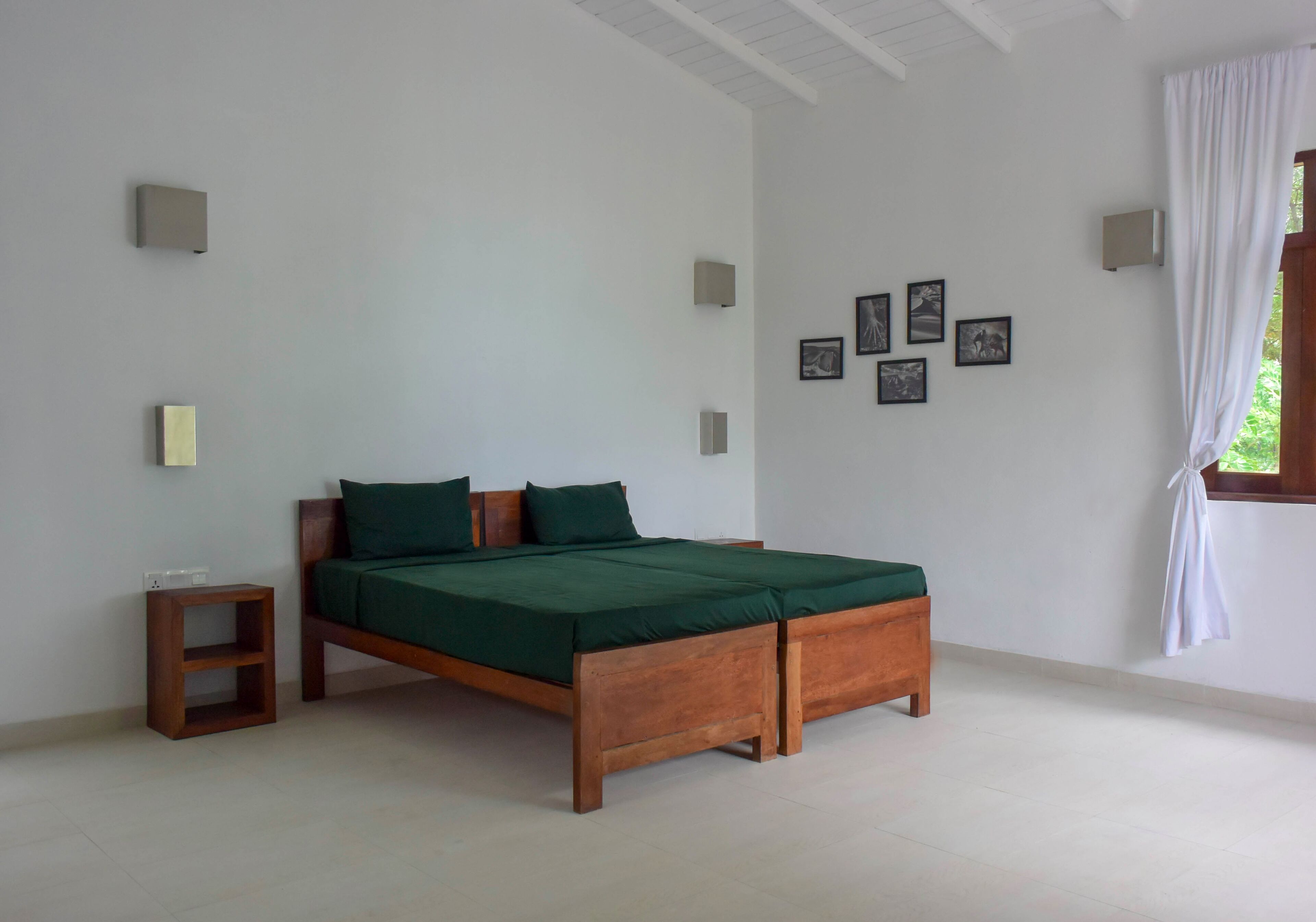 Quarto com cama de madeira, mesa de cabeceira e prateleira pequena. Há luminárias fixadas na parede e quadros emoldurados.