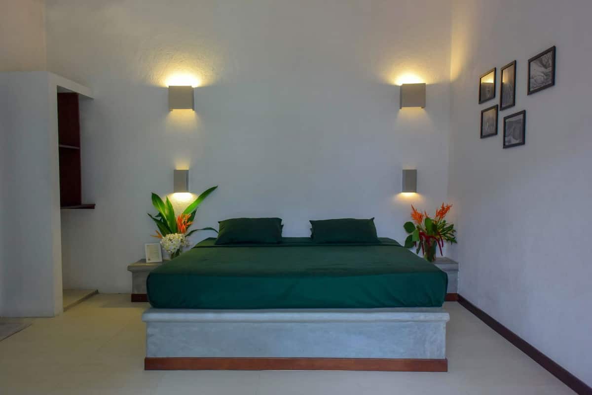 A bedroom with a bed, two bedside tables, a nightstand, a potted plant, and a wall-mounted light fixture.