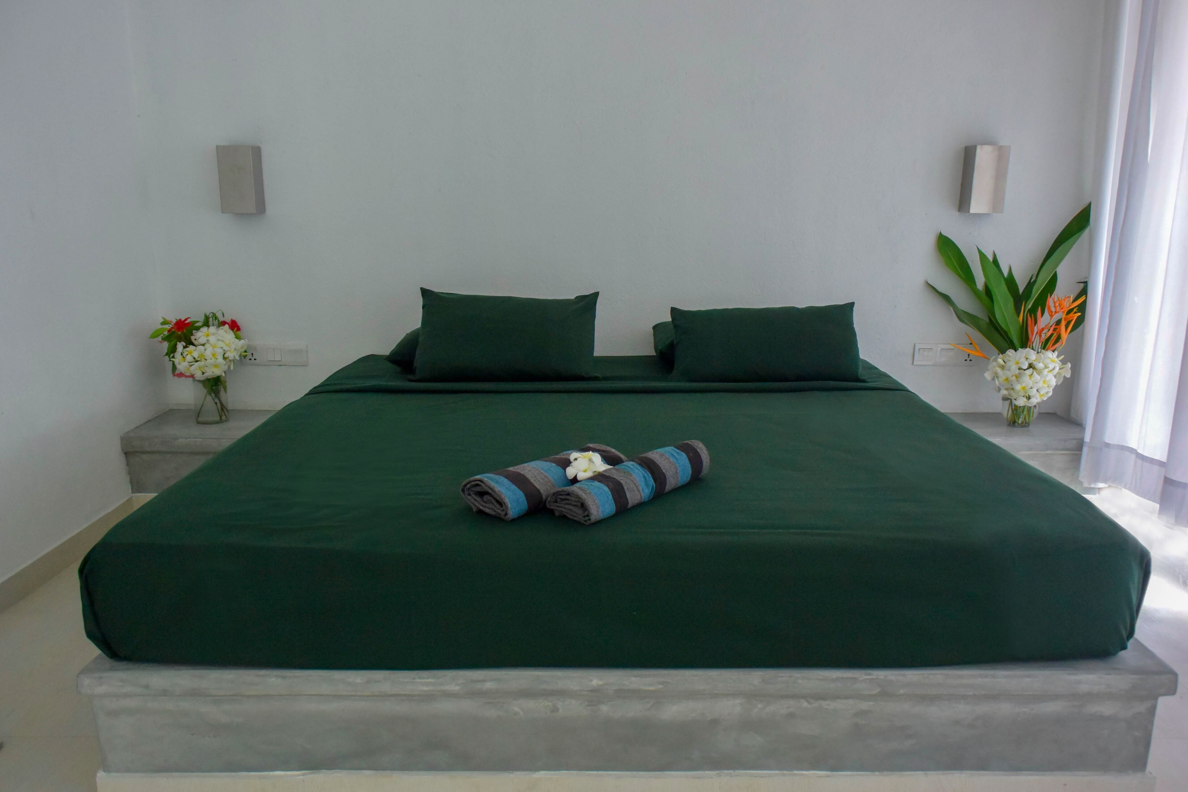 A neatly made bed with a green bedspread and pillows, a rolled towel, and a vase of flowers on a nightstand.