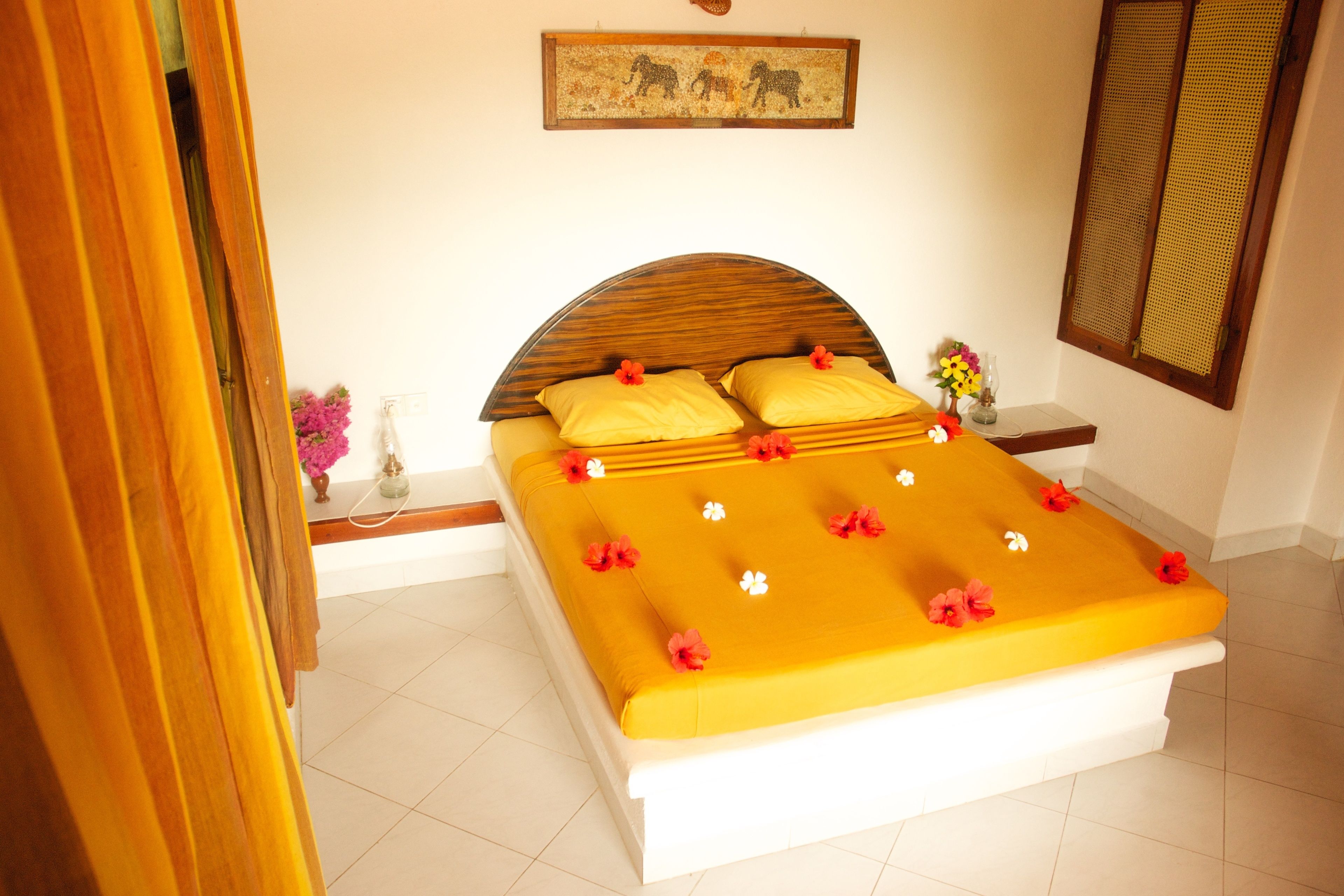 A neatly made bed with a yellow bedspread and red flowers, a wooden headboard, a small bedside table with a vase of flowers, and a wall-mounted picture of elephants.