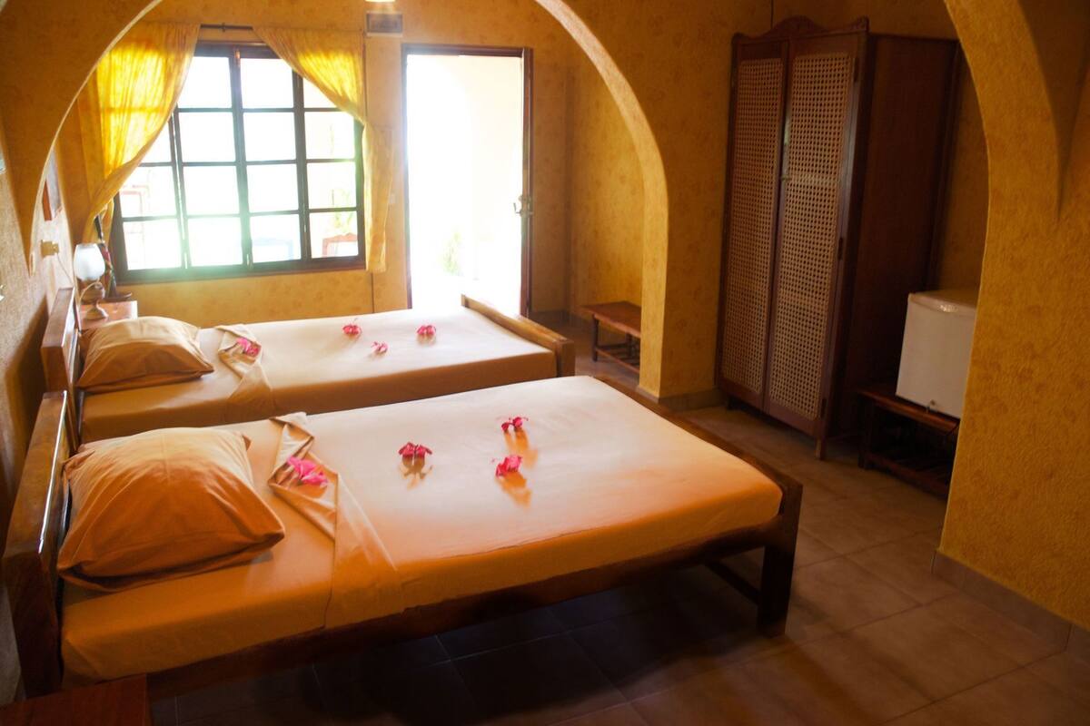 A room with two beds, a window with curtains, a wooden wardrobe, and a small table.