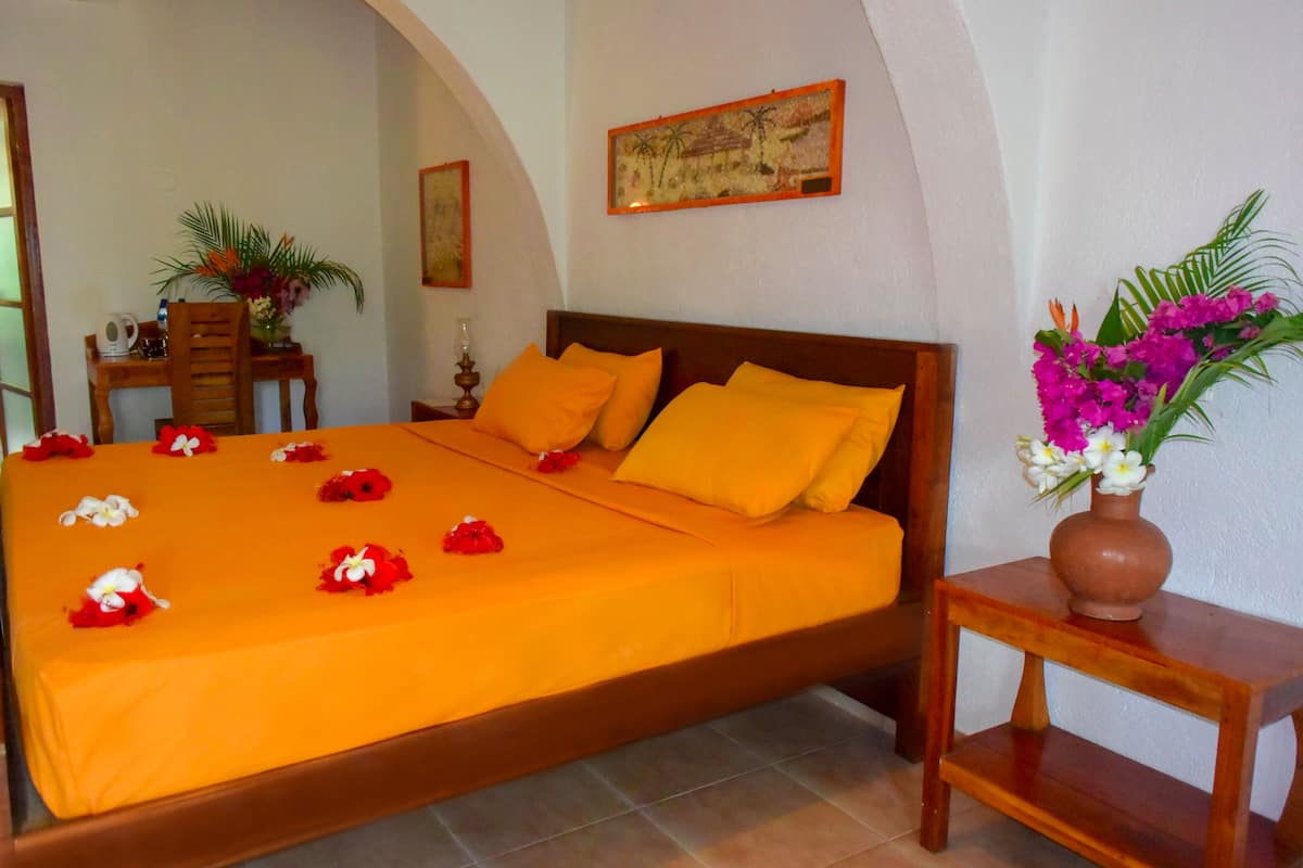 A neatly made bed with orange bedding and pillows, a wooden nightstand with a vase of flowers, and a small table with a vase of flowers.