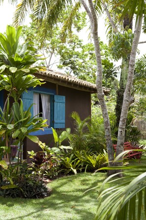 A small house with a tiled roof and blue shutters, surrounded by lush greenery and palm trees. - Casas da Vila (Porto Seguro)