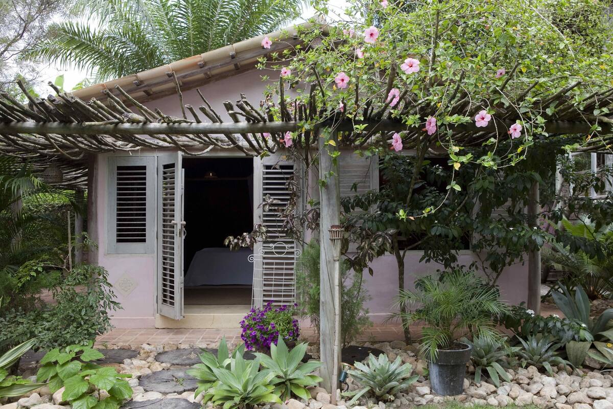 Ein Haus mit einer Holzpergola, die mit rosa Blüten bedeckt ist, ein kleiner Garten mit verschiedenen Pflanzen und eine Tür mit Fensterläden.