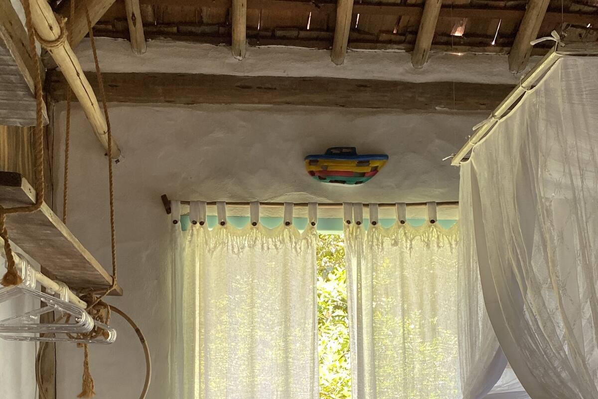 Ein rustikaler Raum mit einem Bett, einem kleinen Tisch, einem Fenster mit durchsichtigen Vorhängen und einem gemusterten Teppich.