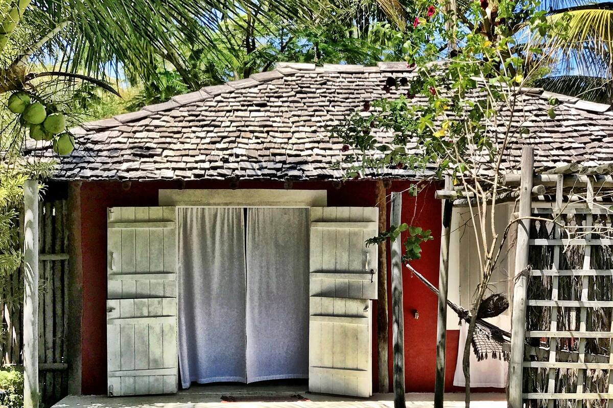 Ein kleines, rustikales Haus mit Ziegeldach, Holztür und einem kleinen Garten mit Rasen und einer Palme.