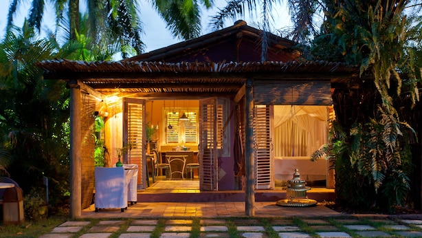 A cozy, well-lit outdoor dining area with a table and chairs, surrounded by lush greenery and a stone pathway leading to a building with a tiled roof. - Casas da Vila (Porto Seguro)