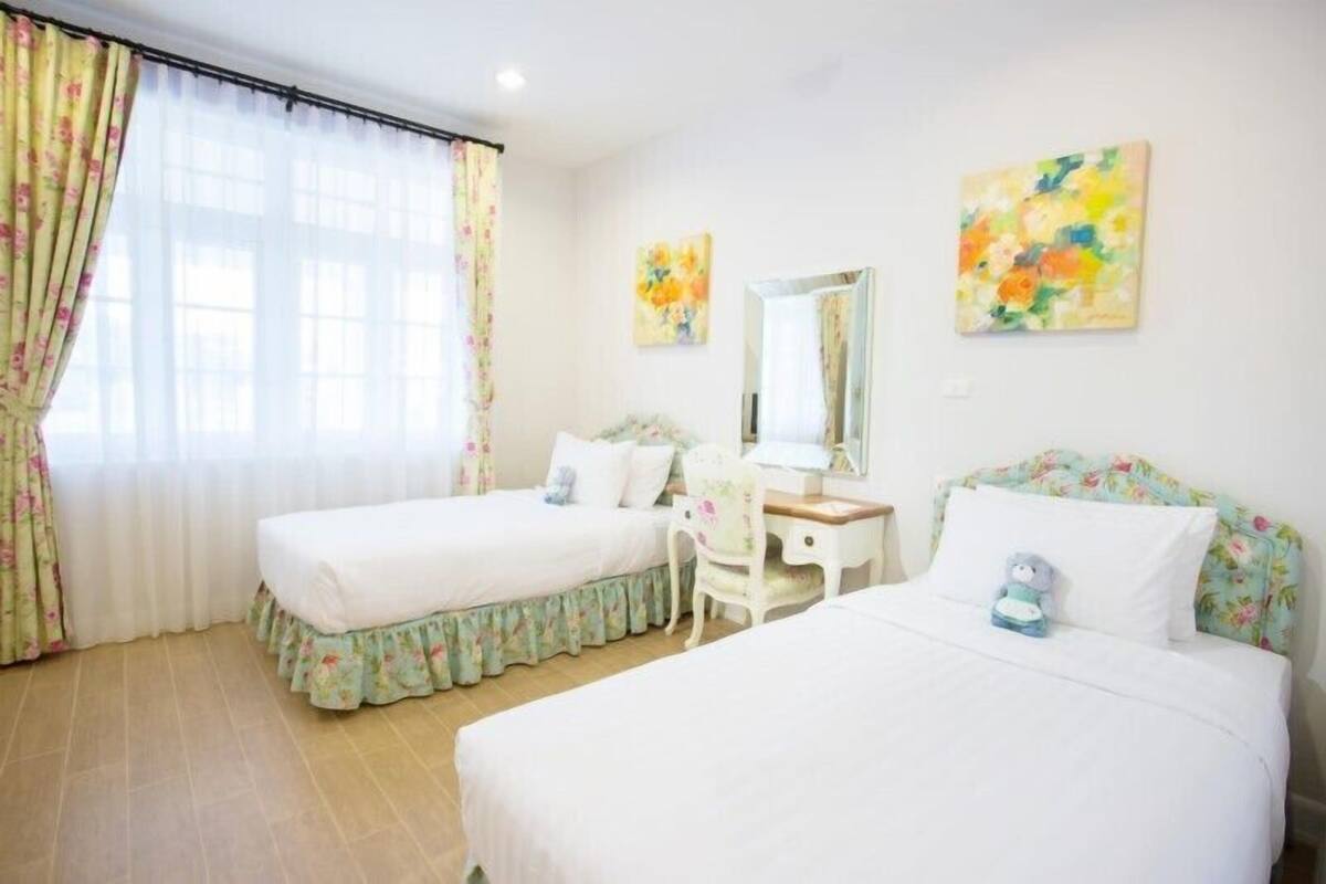 A hotel room with two beds, a desk, a chair, a mirror, and two floral paintings on the walls.
