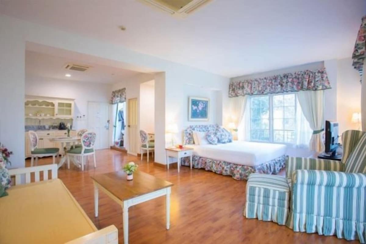A cozy room with a bed, a small table, and a chair. There is a window with curtains and a view of the outside.