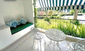 Ein Balkon mit einer gepolsterten grünen Bank, einem weißen Tisch mit aufwendiger Gestaltung und Blick auf einen Garten mit gestreiftem Sonnensegel.