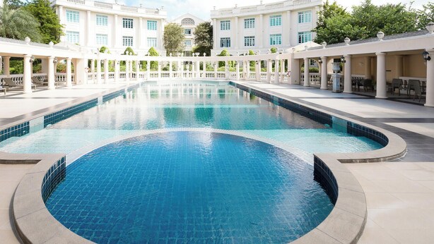 A large, rectangular swimming pool with a tiled edge, surrounded by a white colonnade and a building with multiple windows in the background. - Kensington English Garden Resort Khaoyai (Pak Chong)