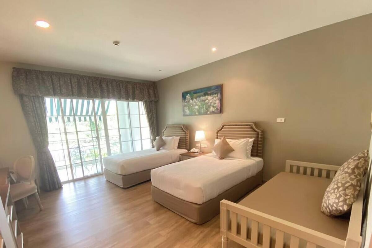 A hotel room with two beds, a small table, a chair, a window with curtains, and a painting on the wall.