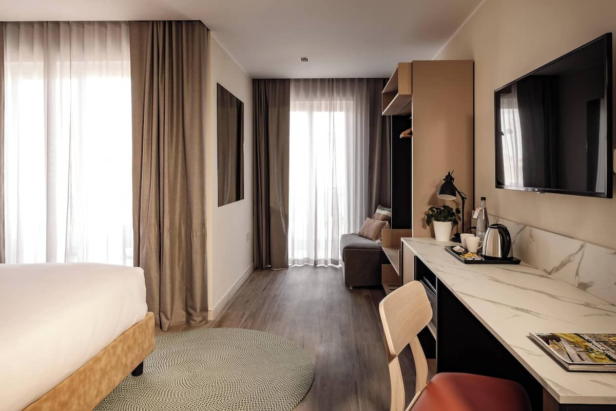 Habitación de hotel con cama, escritorio, silla, televisión y ventana con cortinas.