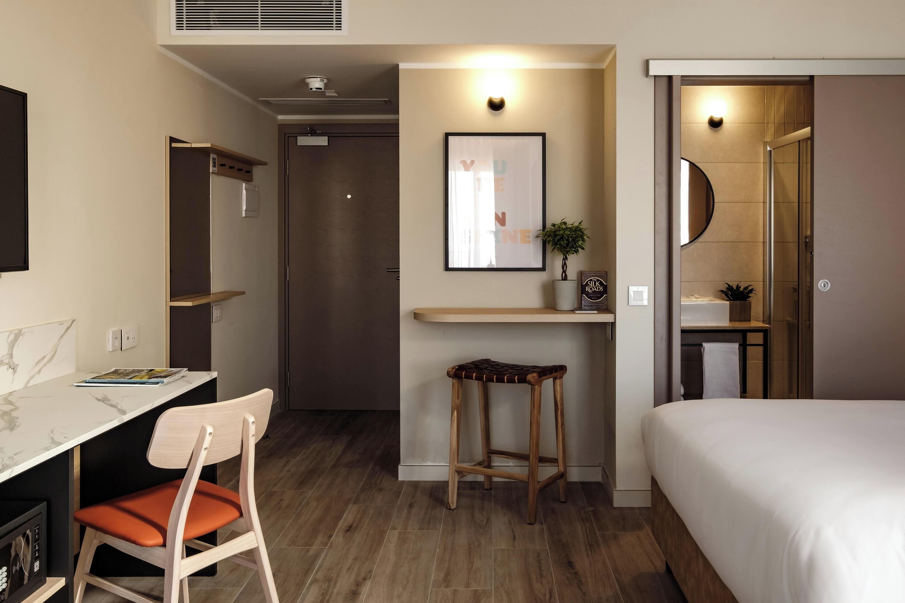 A modern hotel room with a bed, a desk with a chair, a small stool, a framed picture, and a bathroom visible through an open door.