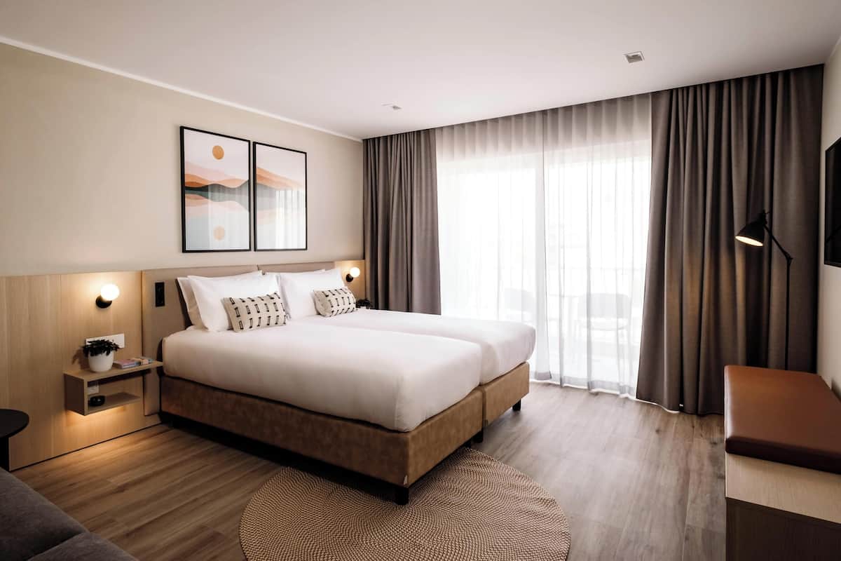 Una habitación de hotel moderna con una cama grande, una mesita de noche con lámpara, una pequeña mesa auxiliar y un amplio ventanal con cortinas.