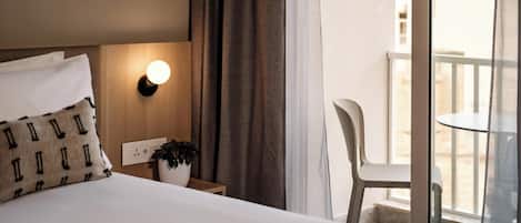 A hotel room with a bed, a nightstand with a lamp, a potted plant, a chair, and a balcony with a railing.