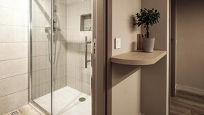 A modern bathroom with a glass-enclosed shower, a wooden shelf with a potted plant, and a light switch on the wall. - Strand Suites By Neu Collective (Gzira)