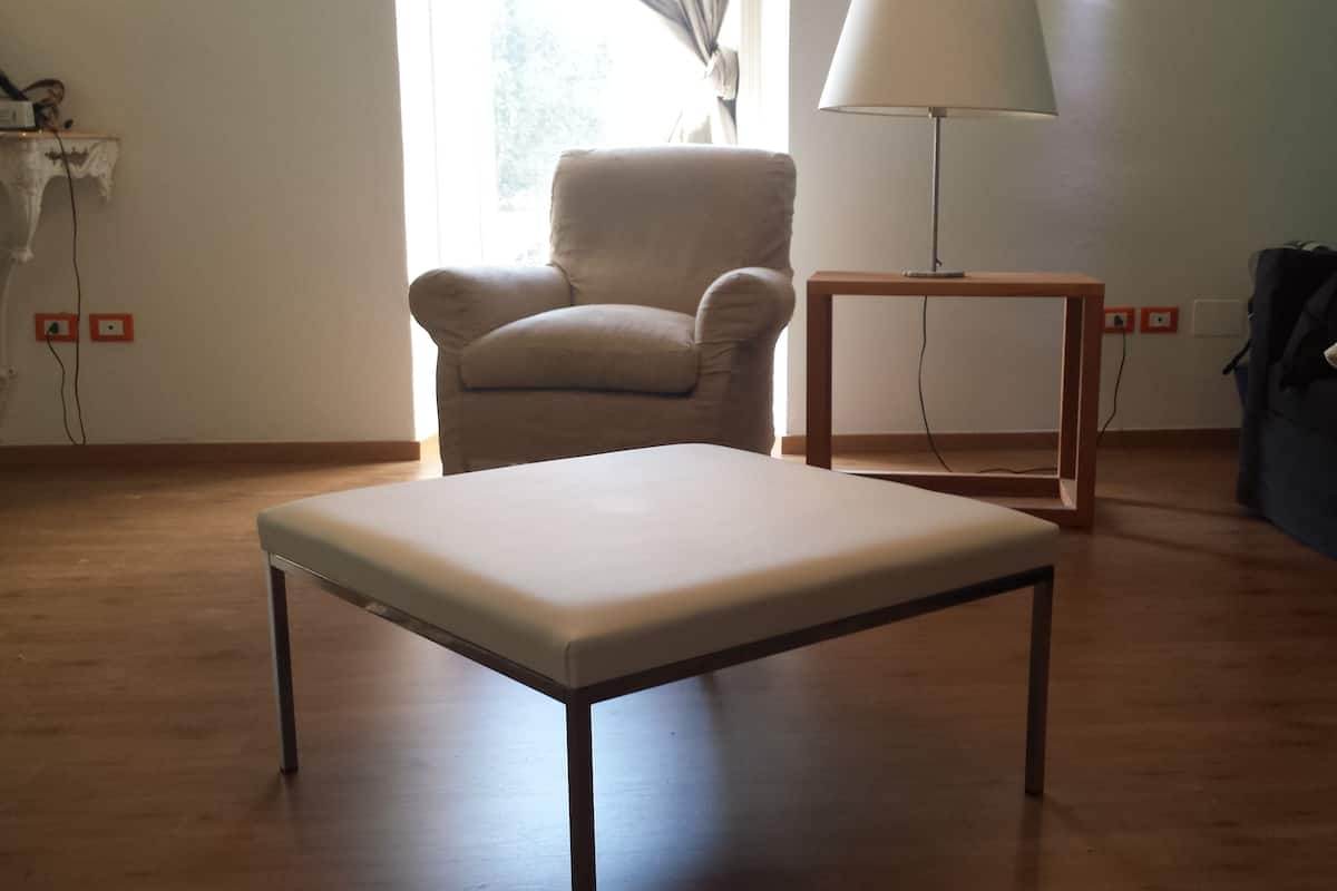 Quarto com piso de madeira, um banquinho branco, uma poltrona bege, uma pequena mesa de madeira com uma luminária e um sofá escuro com um travesseiro.