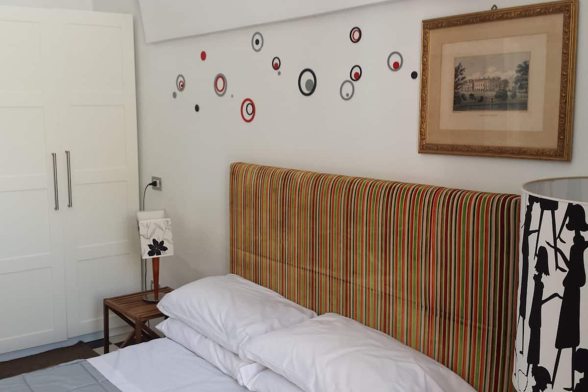 Quarto com cama, cabeceira listrada, guarda-roupa branco, mesinha de cabeceira com abajur e quadro na parede.