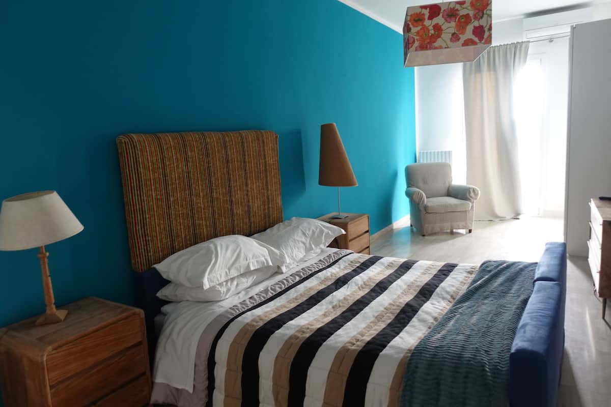 Quarto com cama, mesa de cabeceira, abajur, cadeira e janela com cortinas.