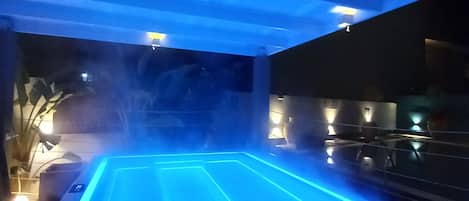 A heated outdoor pool area with a blue-lit edge, surrounded by plants and under a covered roof.