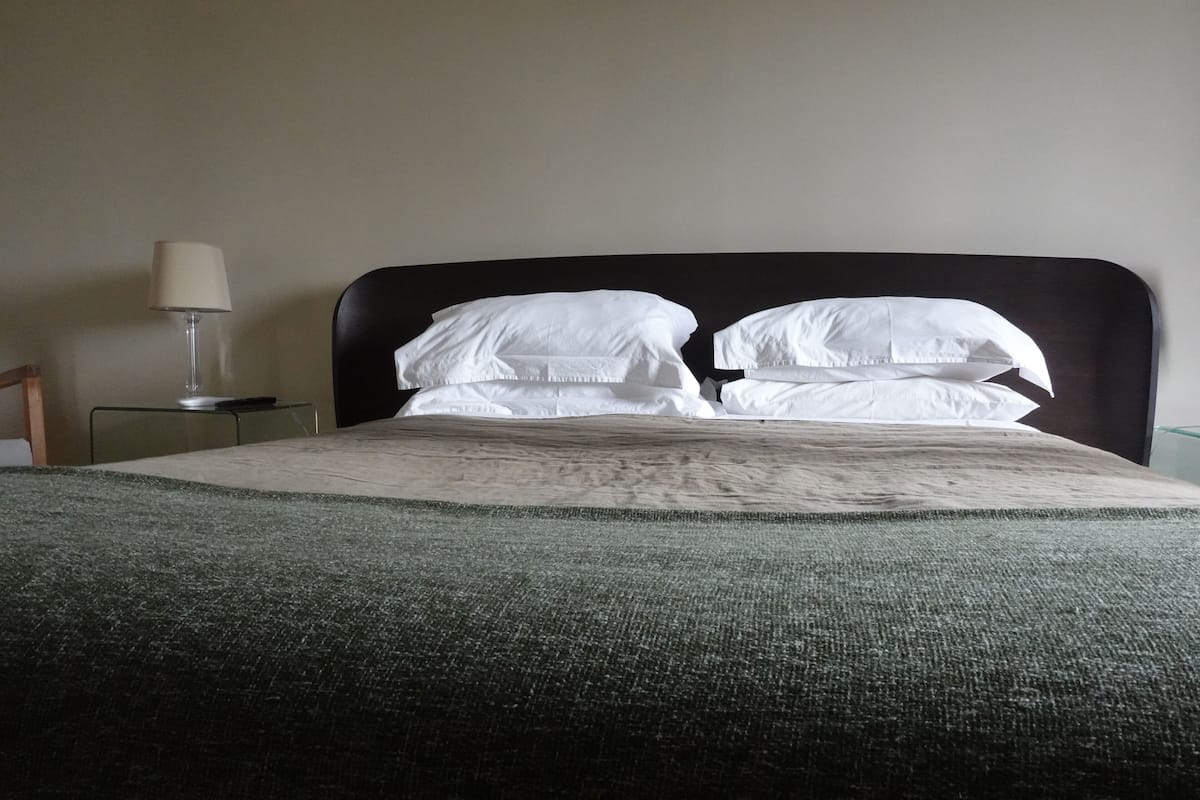 Uma cama bem arrumada com lençóis brancos e um cobertor verde, uma luminária de cabeceira e um painel de cama escuro.