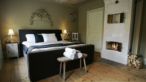 Een slaapkamer met een groot bed, nachtlampen, een klein tafeltje met handdoeken, een open haard en een deur.