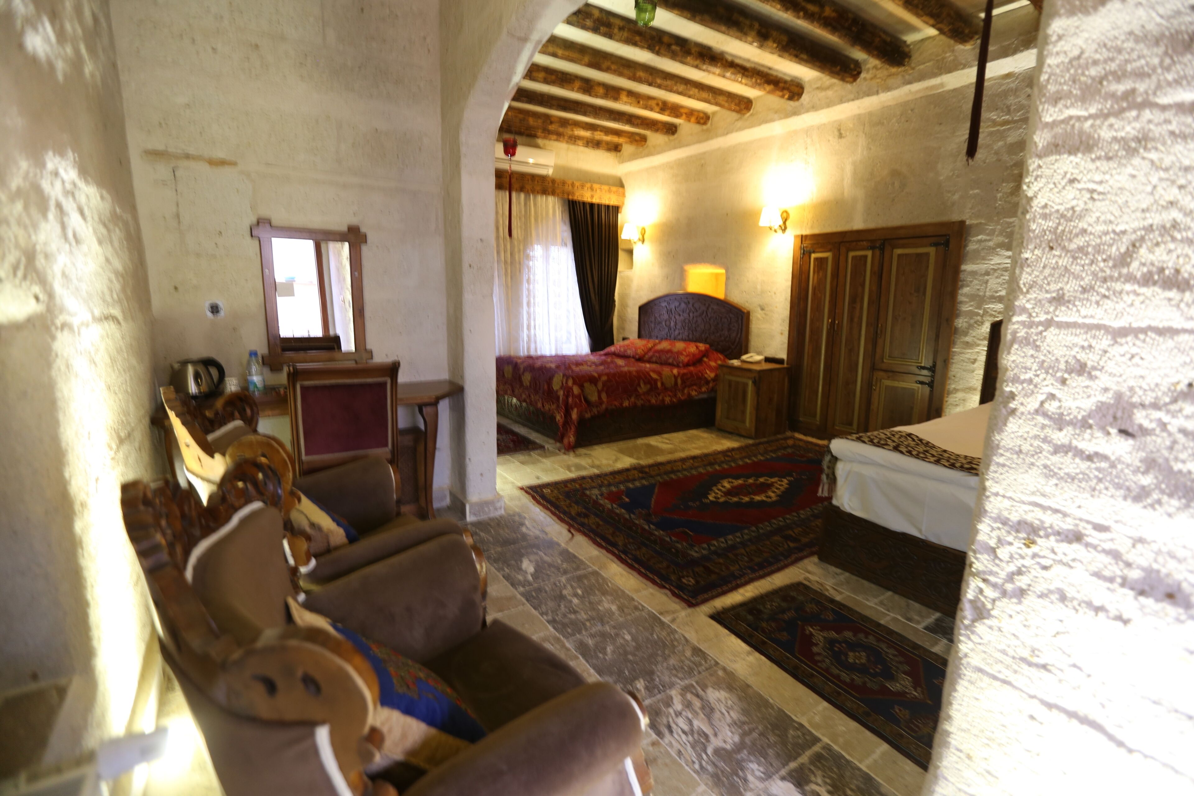 A rustic room with a wooden ceiling, a bed with a red bedspread, a wooden wardrobe, and a small table with a lamp.