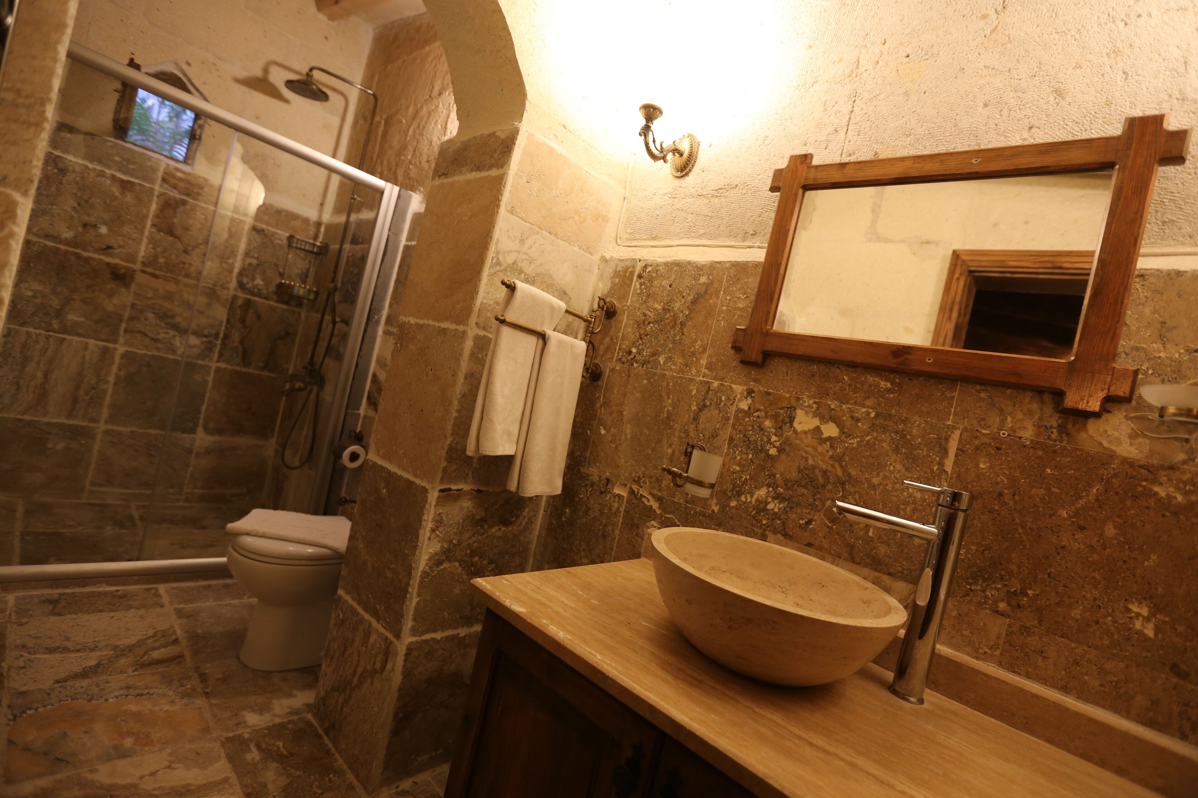 A bathroom with a stone wall, a wooden-framed mirror, a round sink, and a glass shower enclosure.