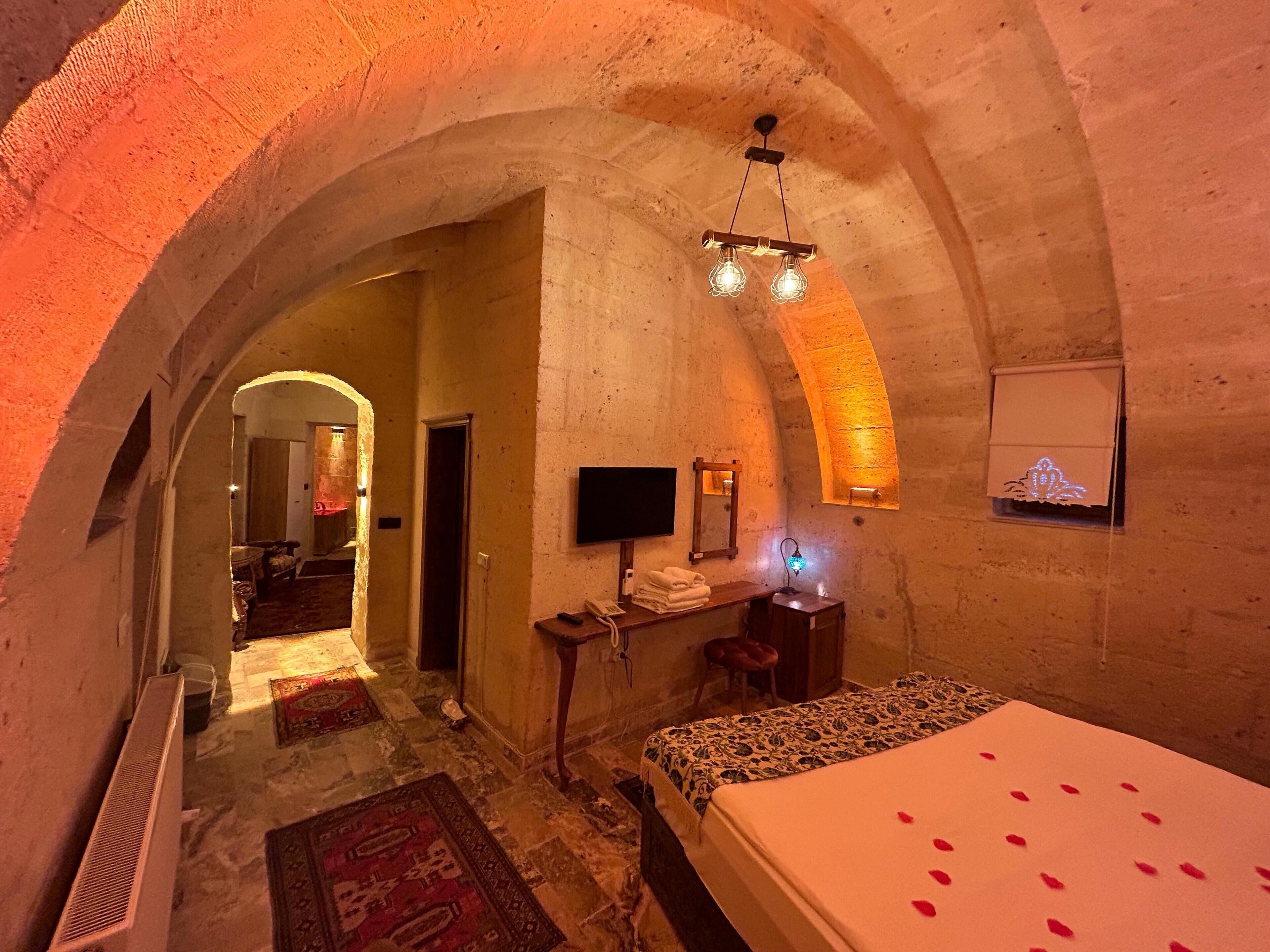 A room with a bed, a desk, and a television. There is an arched doorway leading to another room.