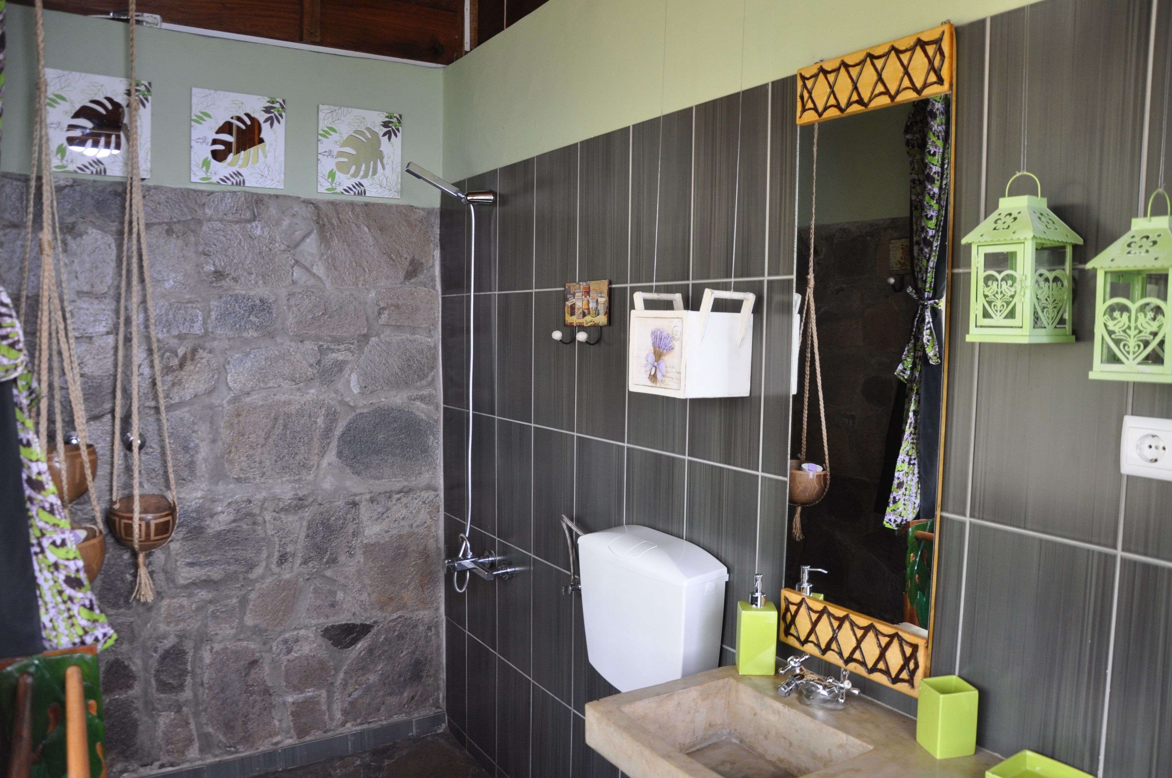 Une salle de bain avec un mur en pierre, une douche, des toilettes, un lavabo et un miroir doté d’un cadre décoratif.