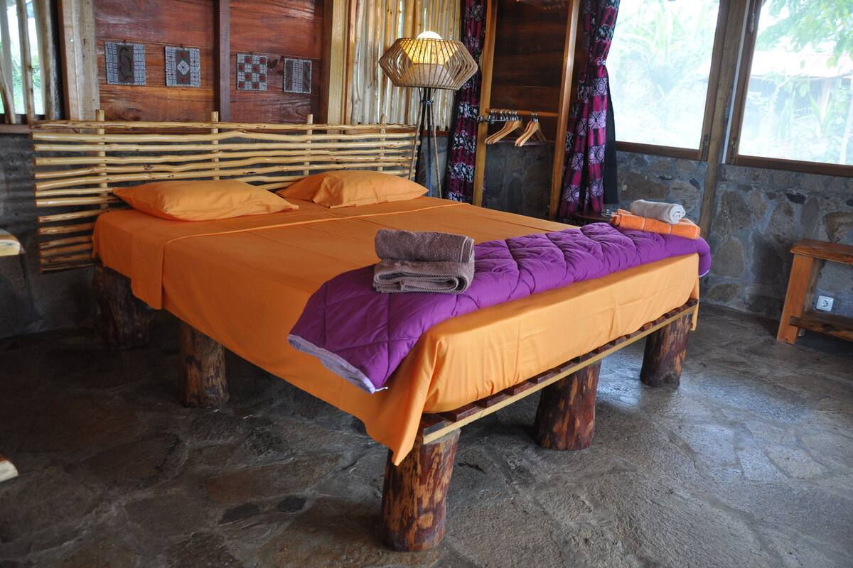 A bed with a wooden frame and orange bedding, a woven headboard, and a stone wall.