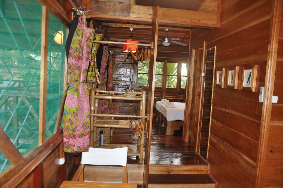 A wooden cabin with a bed, a table, and a chair. There is a window with a view of greenery outside.