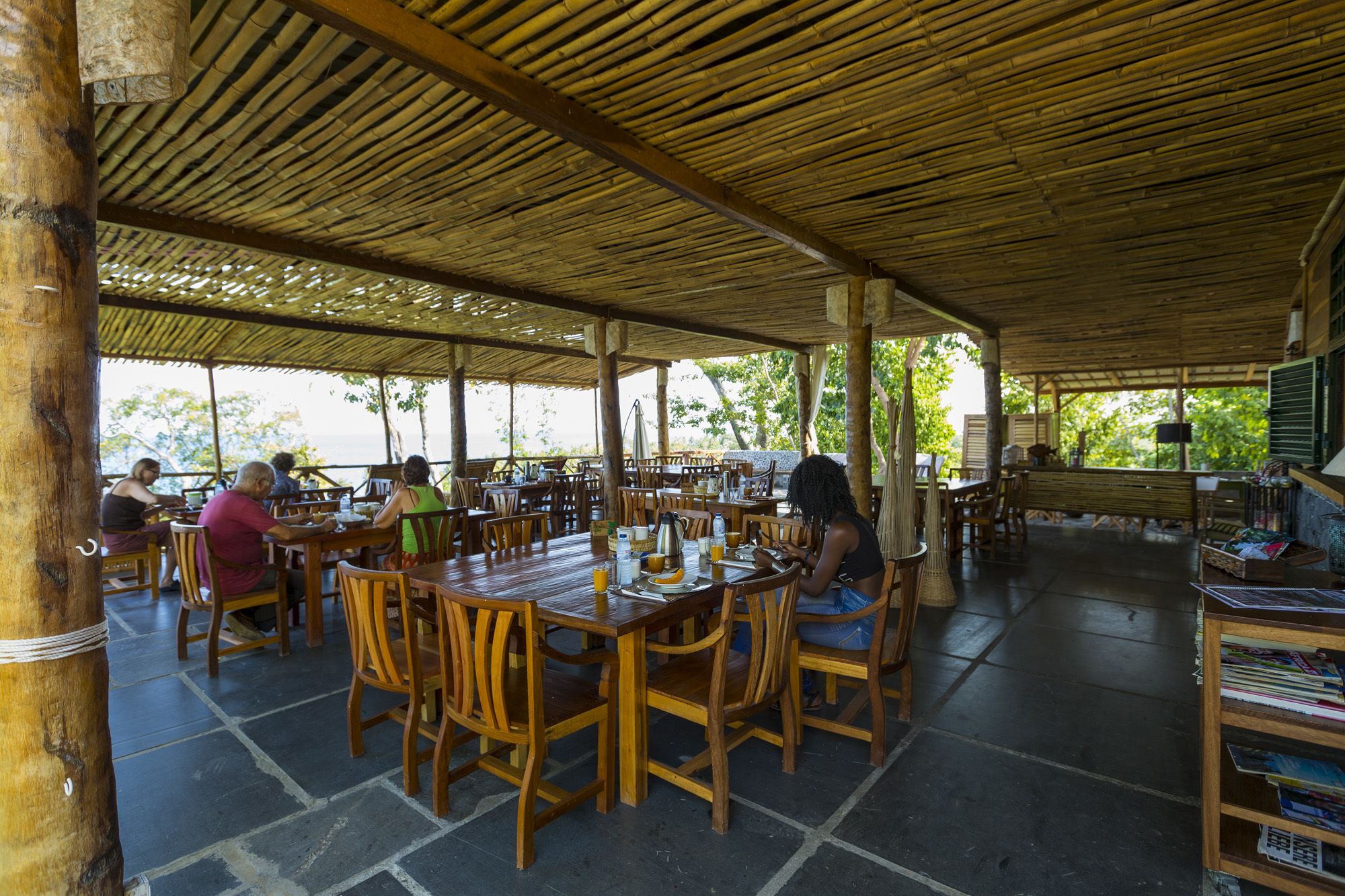 An outdoor dining area with wooden tables and chairs, covered by a bamboo roof, and a view of the sea in the background.