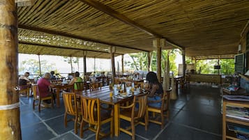 An outdoor dining area with wooden tables and chairs, covered by a bamboo roof, and a view of the sea in the background.