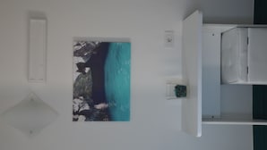 A room with a large painting of a coastal landscape, a white bedside table, and a white bed.