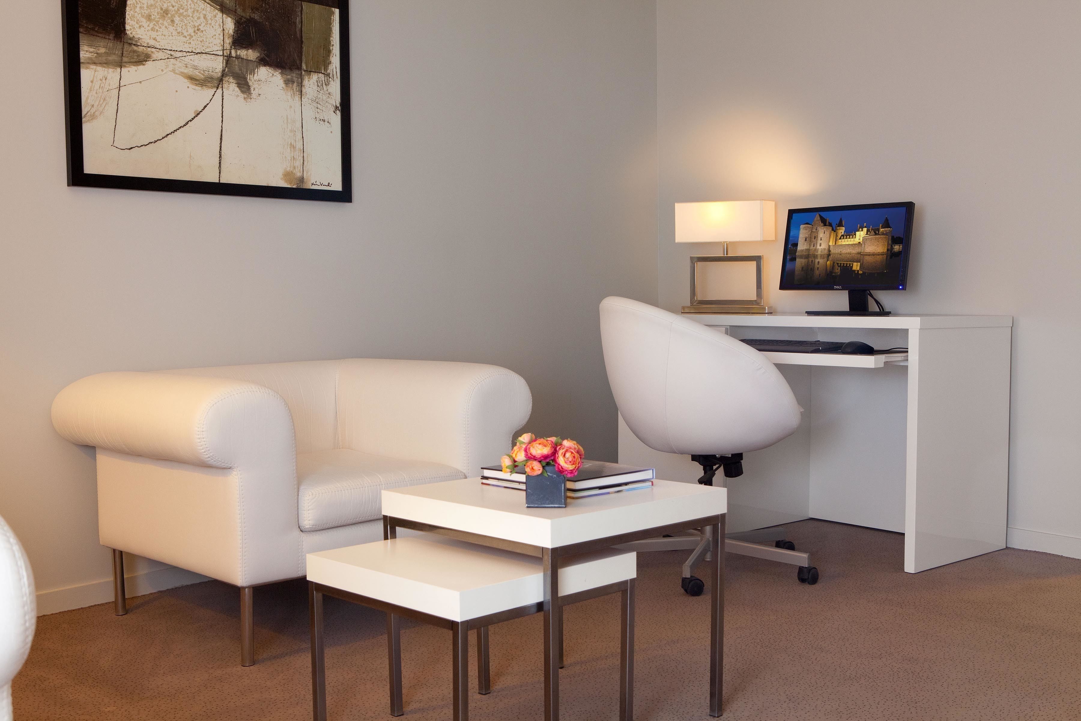 Ein modernes Wohnzimmer mit einem weißen Sofa, einem weißen Sessel, einem kleinen weißen Tisch mit einer Lampe und einem Fernsehgerät auf einem weißen Schrank.