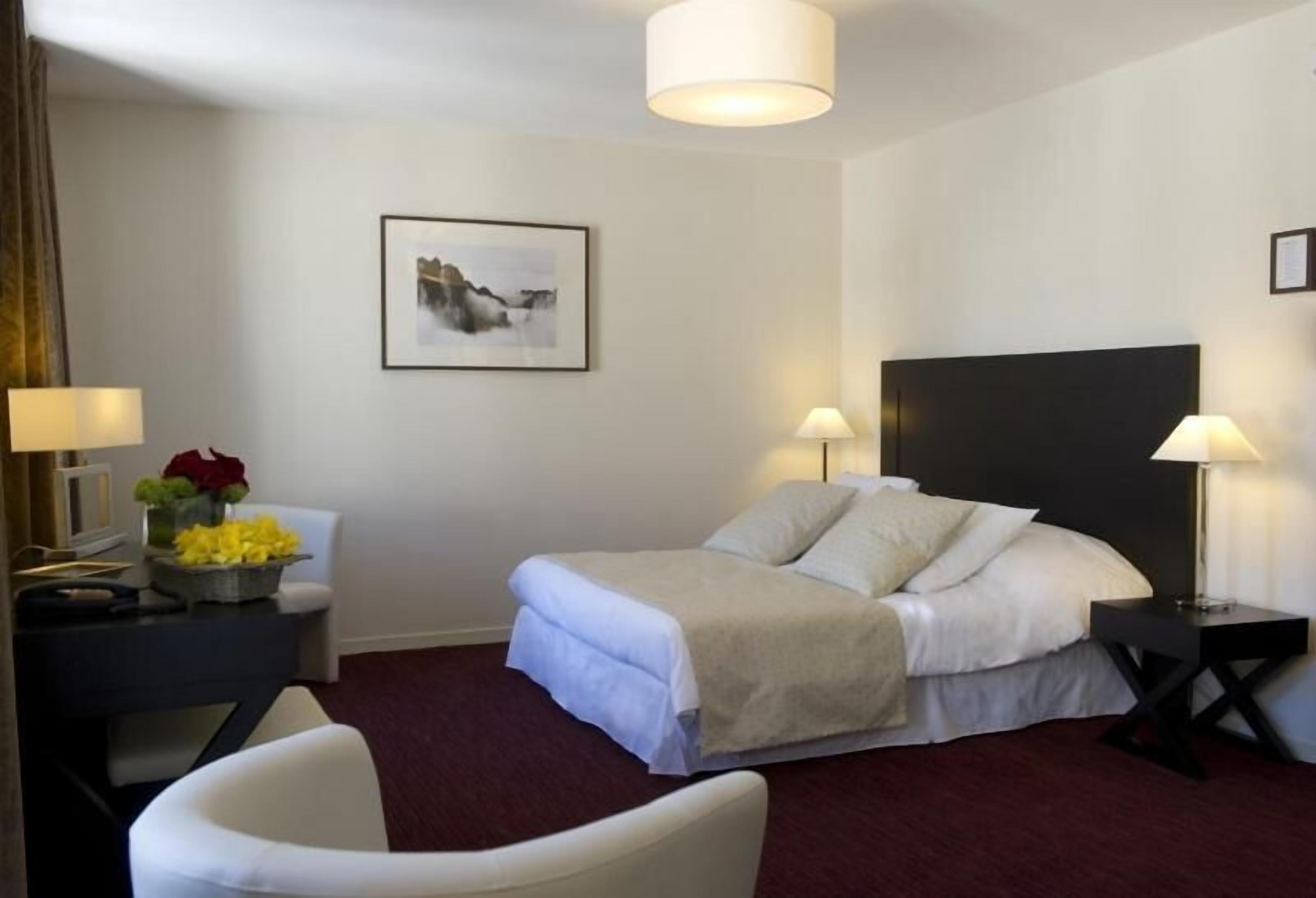 A hotel room with a bed, two bedside lamps, a desk with a lamp, a chair, a framed picture on the wall, and a vase with flowers.