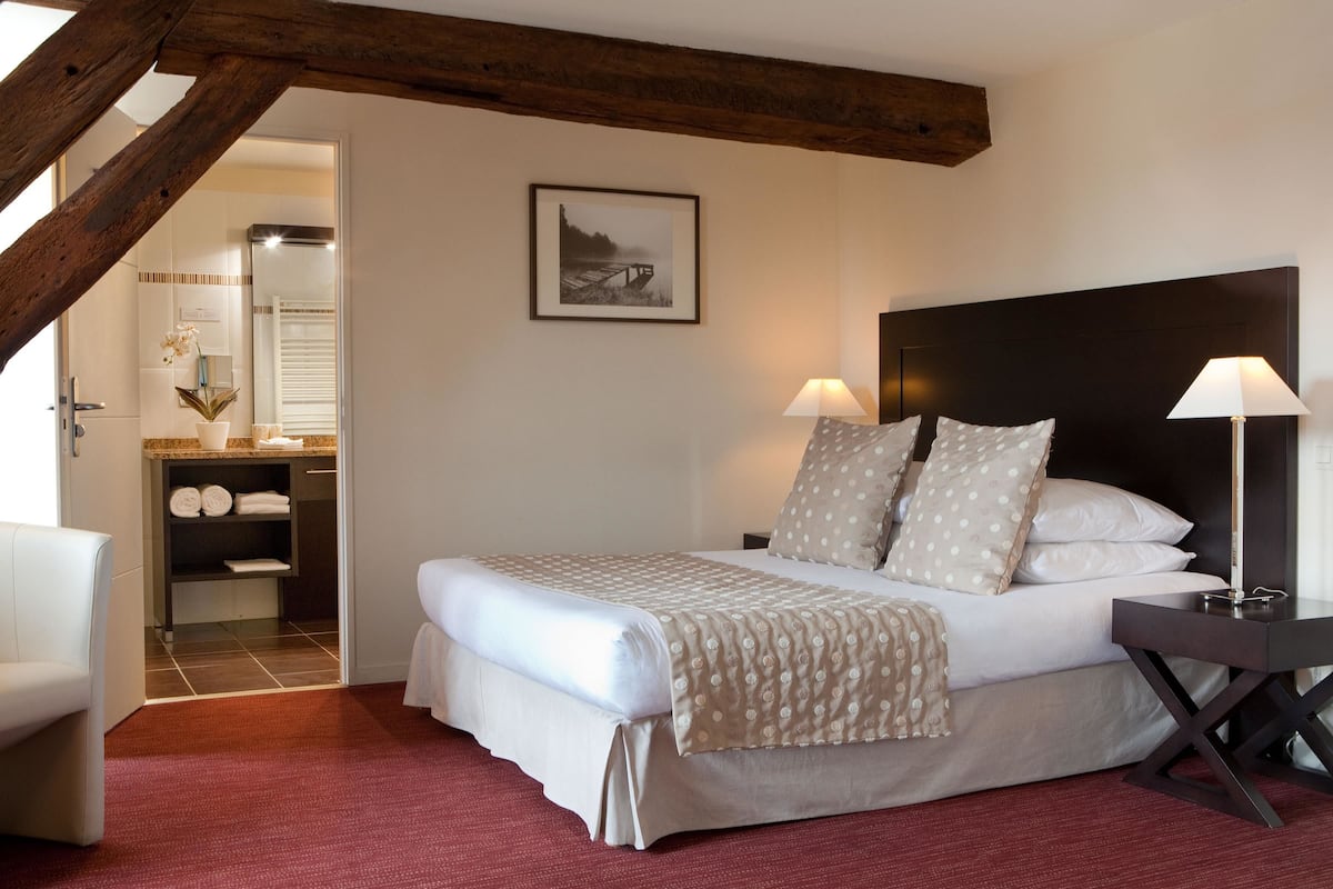 A neatly arranged bedroom with a bed, two bedside tables with lamps, a wooden headboard, a framed picture on the wall, and a bathroom visible through an open door.