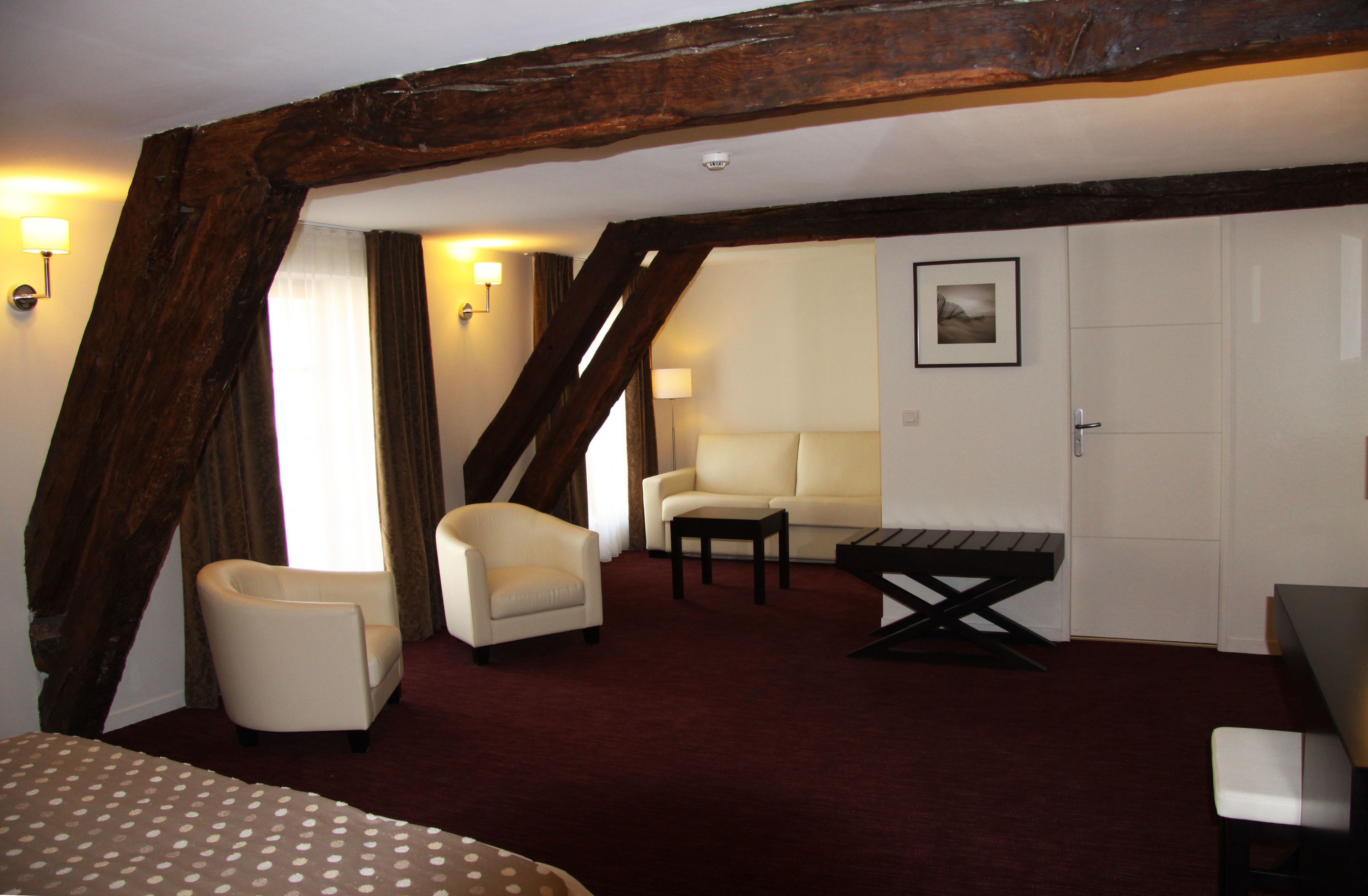 A room with a high ceiling, exposed wooden beams, a sofa, two armchairs, a small table, and a bed with a polka dot bedspread.