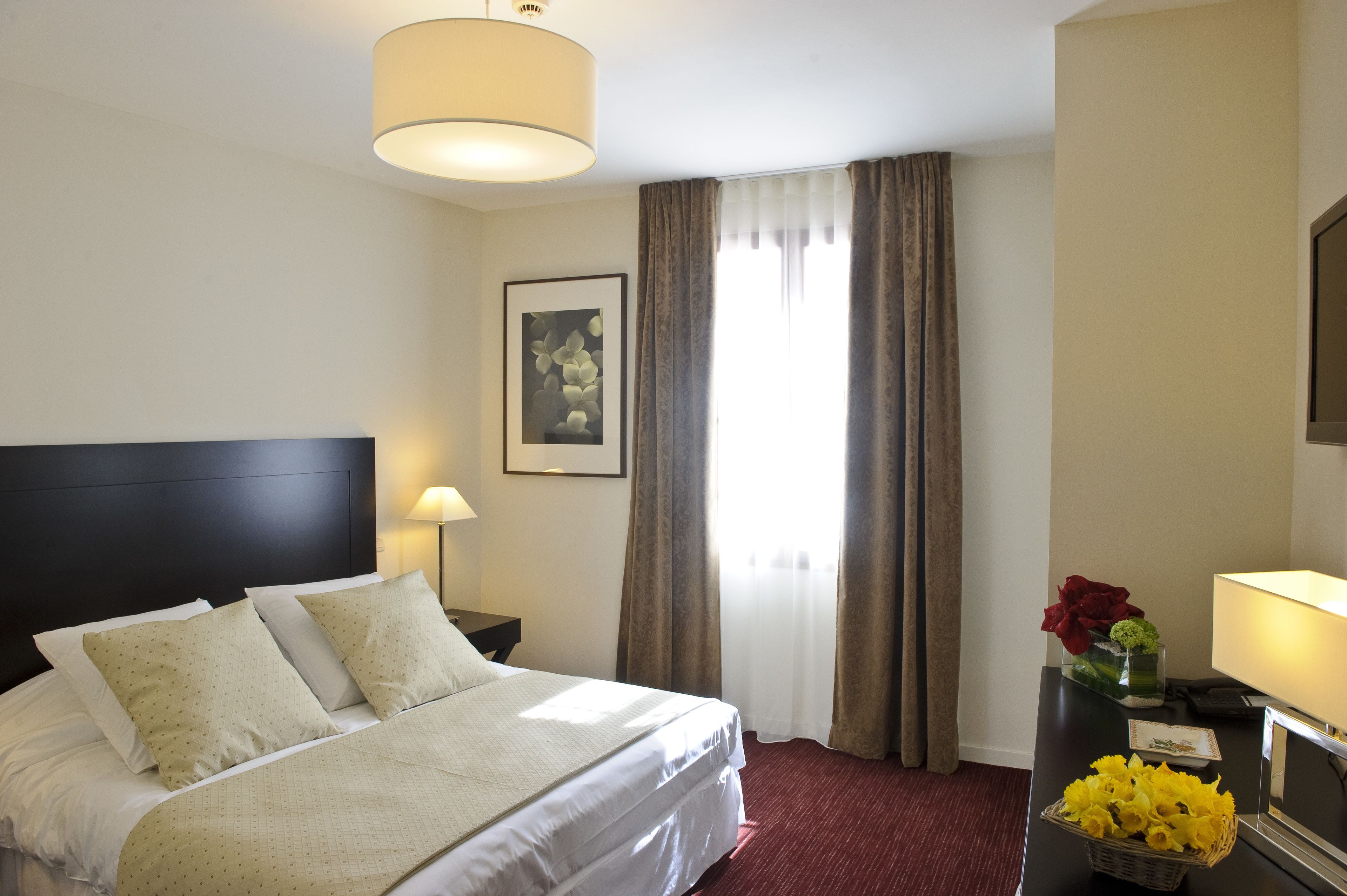 Une chambre d’hôtel avec un grand lit, une table de chevet, une lampe, un tableau encadré au mur et une fenêtre avec des rideaux.